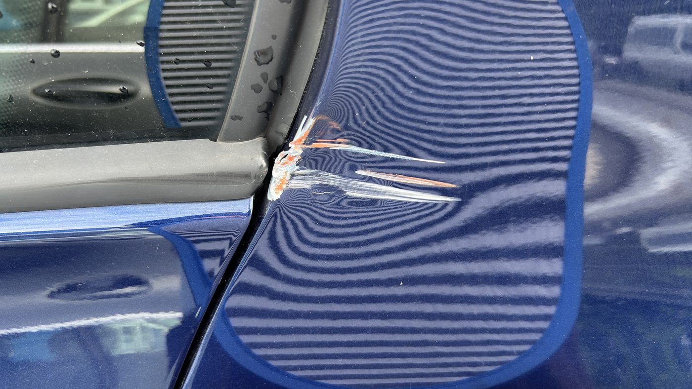 Aufgerissene Naht am Seitenteil hinten links, im Riss der orange Lack des Unfallgegners